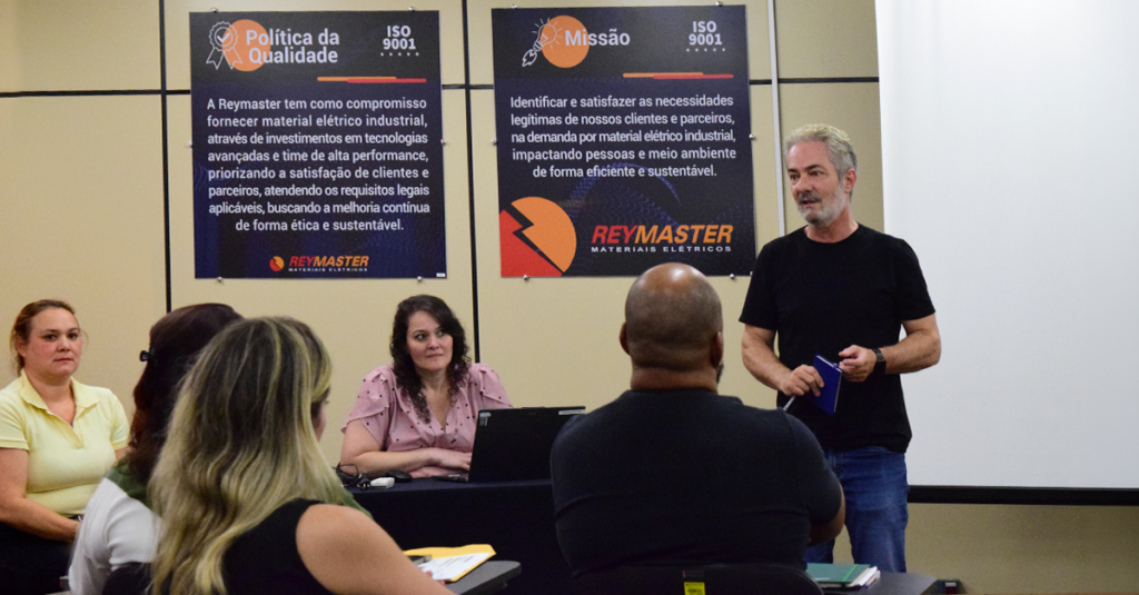 Reunião interna Reymaster com painéis de Política da Qualidade e Missão ISO 9001 visíveis ao fundo